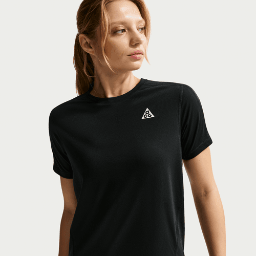 En kvinde med lysebrunt hår i hestehale bærer Nike ACG Short-Sleeve Running Top til kvinder, sort/summit white, med et lille hvidt trekantlogo på brystet - perfekt til trailløb og vist på en neutral baggrund.