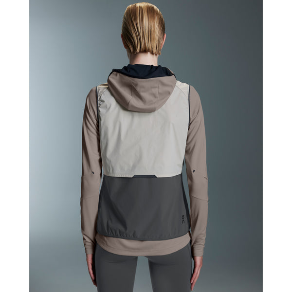 En kvinde med kort, tilbagestrøget blondt hår kigger væk og er iført On Weather Vest Dame in Desert/Eclipse - en elegant, farveblokeret løbevest dame - i beige, grå og creme over grå leggings.