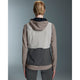 En kvinde med kort, tilbagestrøget blondt hår kigger væk og er iført On Weather Vest Dame in Desert/Eclipse - en elegant, farveblokeret løbevest dame - i beige, grå og creme over grå leggings.