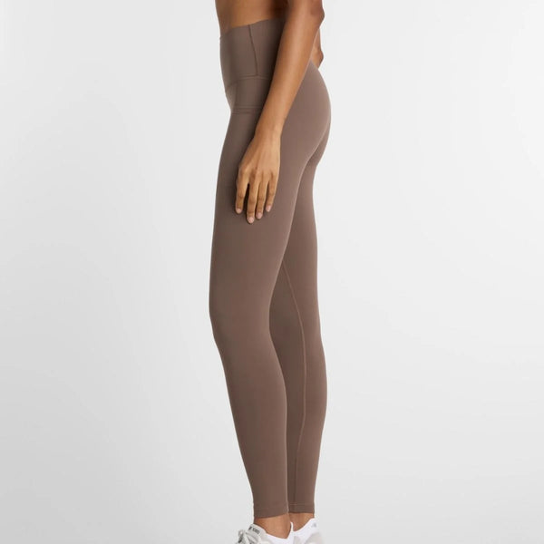 En person står sidelæns i et almindeligt hvidt miljø iført Sleek Pocket High Rise Legging 27, Dame, Cortado og hvide New Balance-sneakers - stilfuld inspiration til træningstøj kvinder. Overkroppen er delvist synlig.