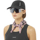 En person iført en Salomon Shakeout Cap (Unisex, Deep Black), store reflekterende solbriller, en sort Salomon-top uden ærmer og et pinkmønstret halstørklæde står på en almindelig hvid baggrund.