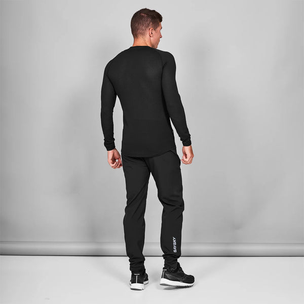 En person kigger væk fra kameraet på en grå baggrund iført Saysky Mesh Base Layer Long Sleeve, Unisex, Black, sammen med sorte bukser med "SWEDEN" på underbenet og sorte sportssko.