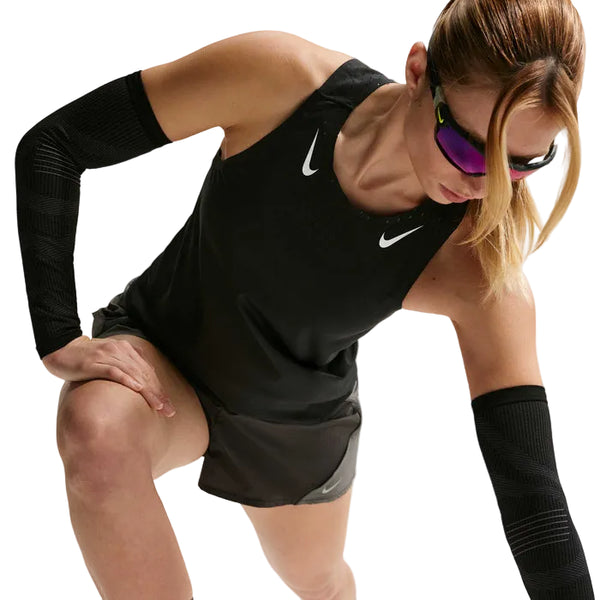 En kvinde i Nike AeroSwift Running Singlet, Dame, Black/White, sammen med sorte shorts, ærmer og solbriller læner sig frem med et bøjet knæ i en atletisk positur, klar til at præstere i åndbart sportstøj.