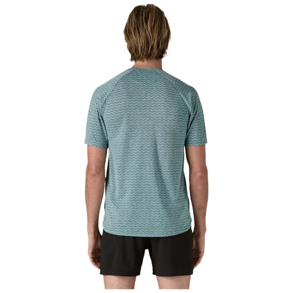 En person med skulderlangt blondt hår står med front mod kameraet iført en Patagonia M's Ridge Flow Shirt, Herre, Wetland Blue sammen med sorte shorts på en almindelig hvid baggrund.
