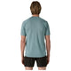 En person med skulderlangt blondt hår står med front mod kameraet iført en Patagonia M's Ridge Flow Shirt, Herre, Wetland Blue sammen med sorte shorts på en almindelig hvid baggrund.