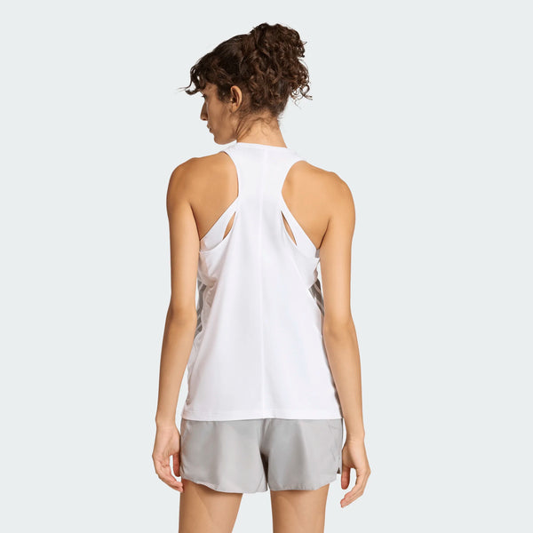 En kvinde med krøllet hår ses bagfra iført en Adidas Iconic Tank Top, Dame, White og lysegrå shorts, stående mod en almindelig lysegrå baggrund.