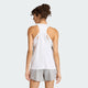 En kvinde med krøllet hår ses bagfra iført en Adidas Iconic Tank Top, Dame, White og lysegrå shorts, stående mod en almindelig lysegrå baggrund.