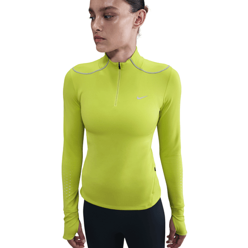 En kvinde i en tætsiddende Nike Therma-FIT ADV 1/2-Zip Reflective Running Top i Cyber/Reflective Silv sammen med sorte leggings står på en almindelig hvid baggrund.