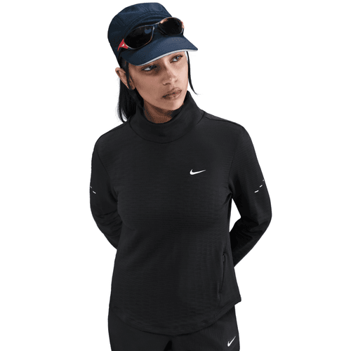 En kvinde står på en hvid baggrund iført Nike Therma-FIT Turtle-Neck Running Top til kvinder i Black/Reflective Silv, sammen med sorte bukser, en marineblå kasket og solbriller, der hviler på hendes kasket.