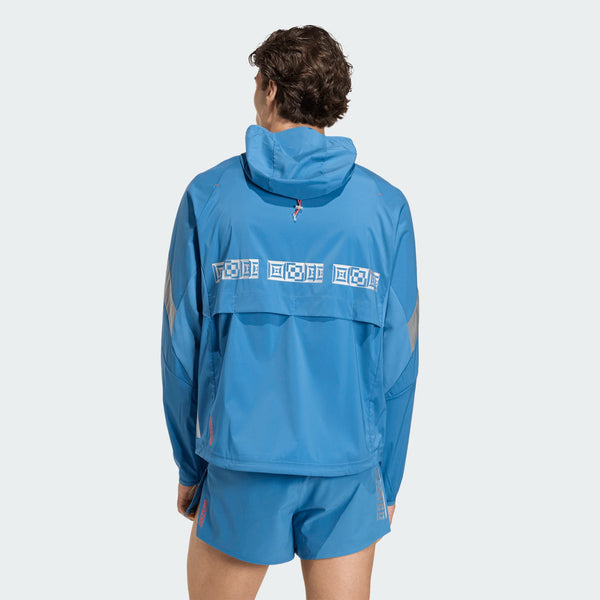 En mand ses bagfra iført en blå Adidas Adizero Hakone Ekiden Jacket (Herre, Duinn/Sliver) med matchende shorts. Den ultralette jakke har en geometrisk mønstret vandret stribe over den øverste del af ryggen mod en lys baggrund.