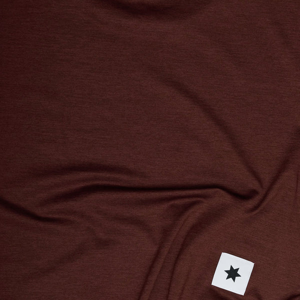 Et stykke rødt stof fra en Saysky W Logo Pace Long Sleeve, Dame, med et hvidt rektangulært mærke med et sort stjernesymbol ses i nederste højre hjørne.
