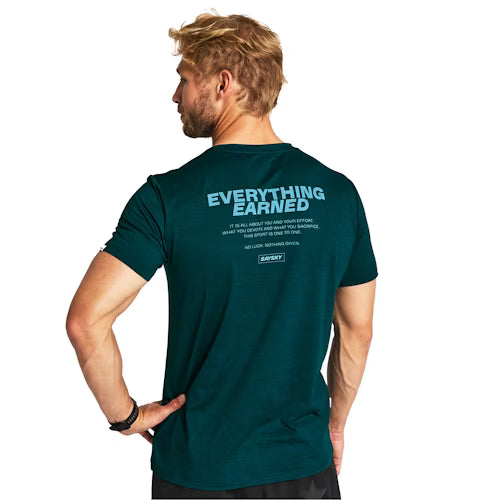 En mand med blondt hår og skæg står med ansigtet vendt væk og er iført en Saysky Statement Pace T-shirt, Herre, Grøn, med blå "EVERYTHING EARNED"-tekst med yderligere mindre tekst på ryggen.