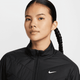 En kvinde med langt sort flettet hår er iført en sort Nike Therma-FIT løbejakke, Dame, Black/Reflective Silv med et hvidt Nike-logo på brystet mod en almindelig lys baggrund.