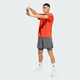 En mand iført Adidas Adi365 Running Tee, Herre, Duor, grå shorts, hvide strømper og hvide sportssko strækker sin højre arm ud over brystet på en lys baggrund - ideel til løbetræning.