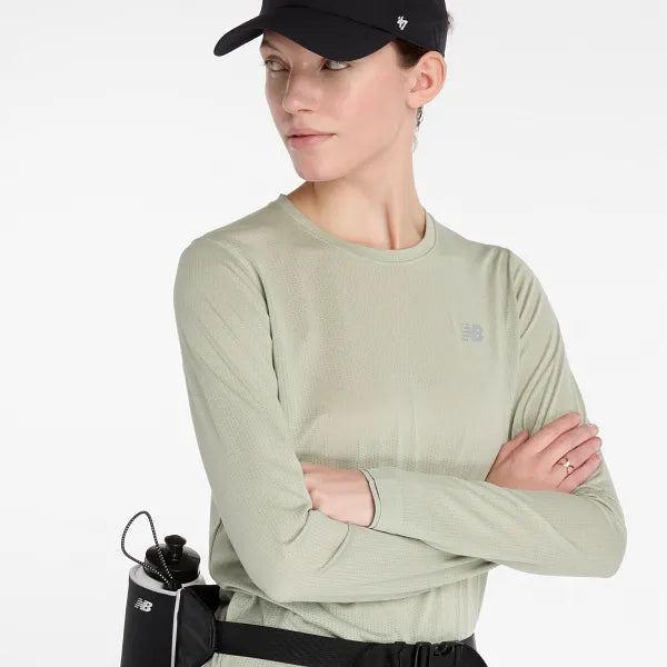 En person iført en New Balance Athletics Long Sleeve Dame shirt i Garter Snake Heather står med armene over kors, parret med en sort kasket og en bæltetaske med en vandflaske og kigger til siden.