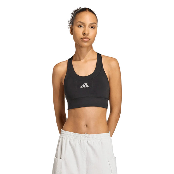 En person står med ansigtet fremad i en Adidas Adi365 Pkt Bra, Dame, Black med hvidt logo og hvide sportsshorts med hænderne på ryggen mod en almindelig lys baggrund.