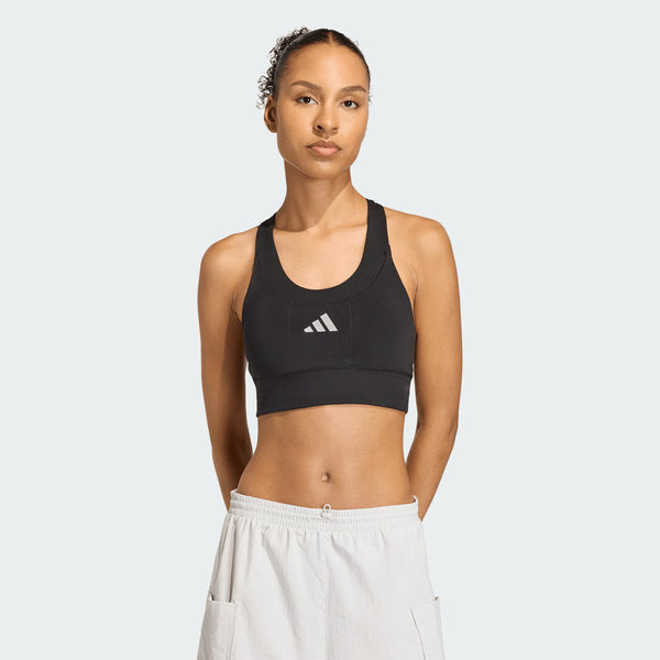 En person står med ansigtet fremad i en Adidas Adi365 Pkt Bra, Dame, Black med hvidt logo og hvide sportsshorts med hænderne på ryggen mod en almindelig lys baggrund.