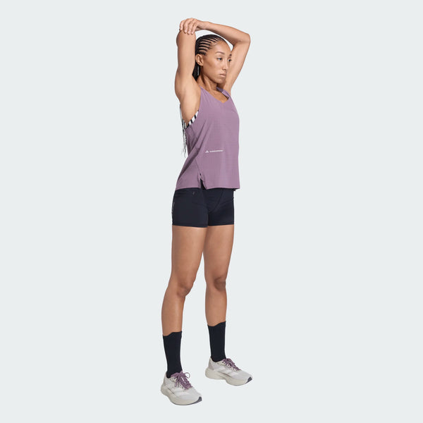 En person står oprejst og strækker sin venstre arm over hovedet. De har en ærmeløs lilla top på, sorte shorts over Adidas Adi365 Hk Tight, Dame, sorte strømpebukser med høj talje, sorte sokker, hvide sportssko. Baggrunden er almindelig lysegrå.