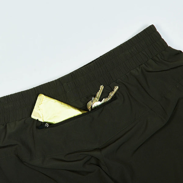 Nærbillede af en lynlåslomme på grønne Saysky Pace Pants til mænd med nøgler og en gul telefon eller pung på en lys baggrund.