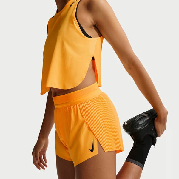 En person i en orange sportstop uden ærmer og Nike AeroSwift Mid-Rise 3In Running Shorts for women i Laser Orange/Black/White/Black strækker benet bag sig og holder foden mod en almindelig lys baggrund.