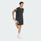 En mand iført sort sportstrøje, Adidas Adi365 Shorts (Herre, Black), hvide sokker og hvide sneakers står på et ben, strækker det andet ben bagud og holder anklen mod en almindelig lys baggrund.