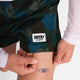 En person bærer blå camomønstrede Flower Pace Shorts 5" fra Saysky med et SAYSKY CPH-mærke og løfter op i kanten for at afsløre en øjenformet lårtatovering. De åndbare shorts til mænd passer perfekt sammen med en hvid langærmet skjorte.