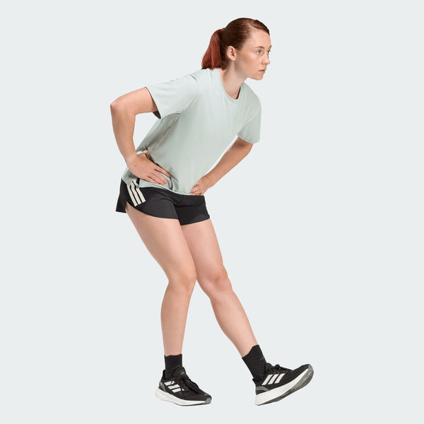 En kvinde i sportstøj bøjer sig frem for at strække sine haser iført en lys t-shirt, sorte Adidas Adi365 Shorts, Dame og sorte sneakers - de perfekte lette shorts til aktive kvinder.