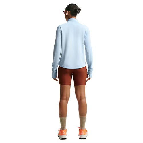 En person står med ryggen til og er iført en Nike ACG Half Zip Running Top til kvinder i Hydrogen Blue/Summit White med rødbrune shorts, lysebrune sokker, orange og hvide sneakers og solbriller med hvidt stel på en almindelig hvid baggrund.