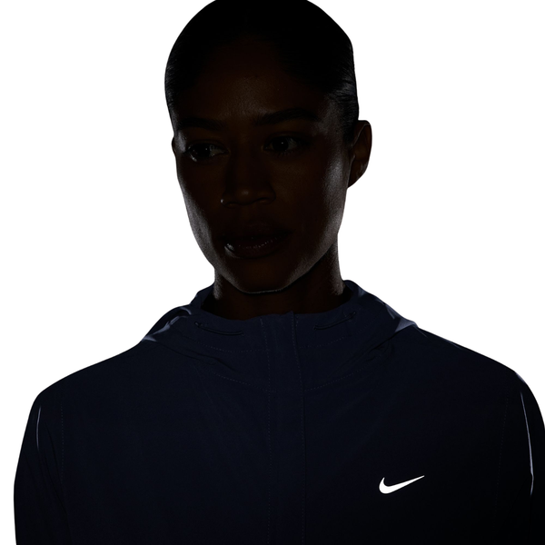 En person iført en mørk lilla Nike Swift Repel Packable løbejakke til kvinder med et synligt, reflekterende sølvfarvet Nike-logo står foran en hvid baggrund; personens ansigt er stort set i skygge.
