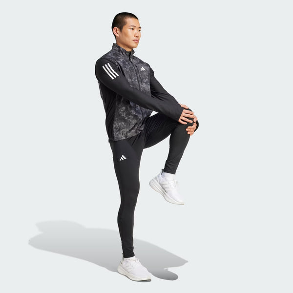 En person i sort Adidas sportsjakke og bukser med hvide striber, hvide sneakers og Adidas Otr Sd Vest M (Herre, Grefou/Black) står på det ene ben og strækker det andet mod en almindelig lys baggrund.