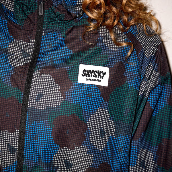 Nærbillede af en person iført Saysky W Flower Pace Jacket, Dame, Blue Aop. Kun den nederste del af ansigtet og det krøllede hår er synligt, med et hvidt SAYSKY COPENHAGEN-mærke på brystet.