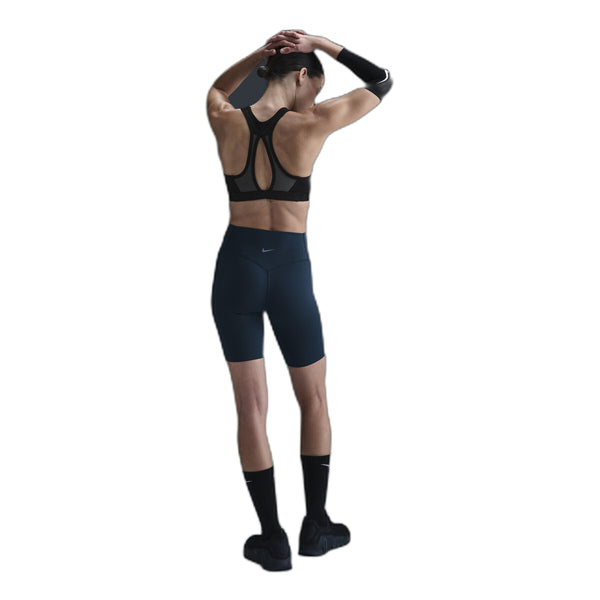 En person iført sort sports-bh, Nike Medium-Support High-Waisted 8" Biker Shorts i Black/Anthracite/Dk Smoke Grey, sorte sokker og sorte sko står med ryggen til kameraet med løftede arme og hænderne foldet bag hovedet.