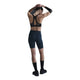 En person iført sort sports-bh, Nike Medium-Support High-Waisted 8" Biker Shorts i Black/Anthracite/Dk Smoke Grey, sorte sokker og sorte sko står med ryggen til kameraet med løftede arme og hænderne foldet bag hovedet.
