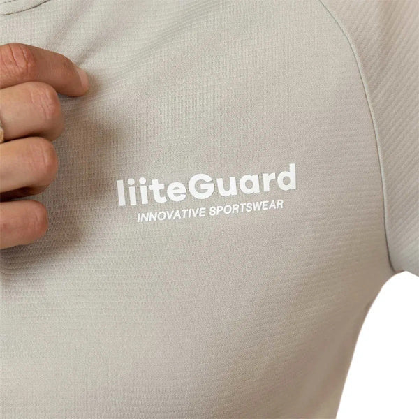 Et nærbillede af en kvinde iført den beige Liiteguard Ground-Tech 1/4 Zip med det hvide INNOVATIVE SPORTSWEAR-logo på brystet. Billedet viser kun en del af hendes hånd og overkrop i denne svedtransporterende træningstrøje dame.