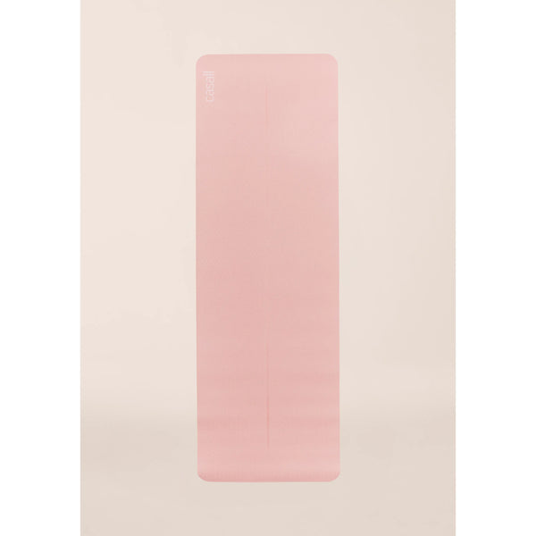 En Casall Yoga Mat Position 4mm, Unisex, i lys rosa/pink med afrundede hjørner ligger fladt på en lys beige baggrund og viser sin glatte, let strukturerede overflade og Casall-logoet i øverste venstre hjørne.
