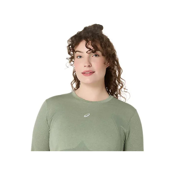 En person med langt krøllet hår bærer Asics Road Seamless Ls Top, Dame i Monument Blue/Whisper Green, med et lille hvidt logo på brystet. Den åndbare træningstop skiller sig ud fra den ensfarvede hvide baggrund.