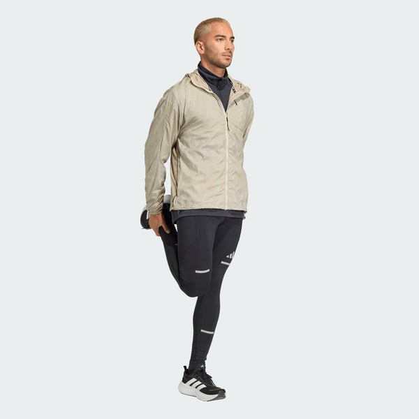 En mand med kort blondt hår bærer en beige Adi365 High-Vis Jacket, Herre, Woca by Adidas, sorte atletiske leggings og hvide sneakers, mens han står på det ene ben og holder den anden fod bag sig i en quadriceps-strækning.