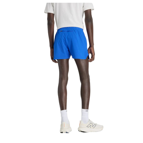 En person kigger væk fra kameraet iført en hvid T-shirt, New Balance RC Short 5" Herre i Blue Gemstone, hvide sokker og råhvide New Balance-sneakers. Baggrunden er helt hvid.