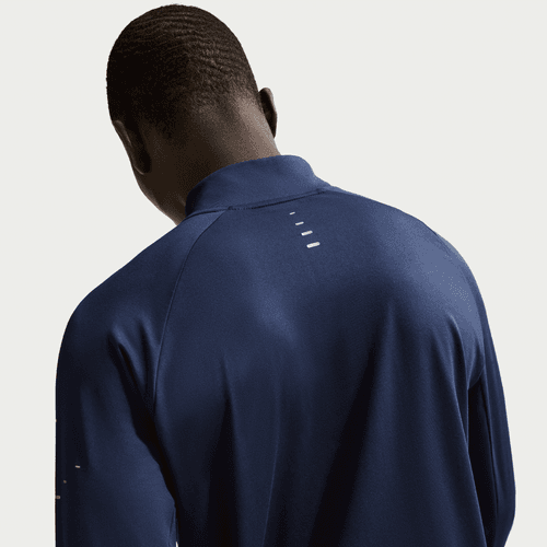 Set bagfra er en person med kort hår iført en Nike Stride Half Zip Midlayer Running Top til mænd i Midnight Navy/Reflective Silver med fire små hvide vandrette linjer på den øverste del af ryggen. Baggrunden er ensfarvet og lys.