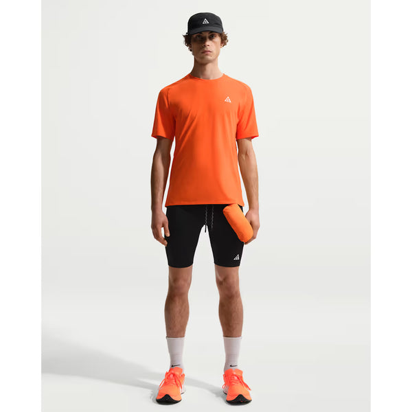 En person kigger fremad iført Nike ACG Dfadv Solar Chase SS til herre i Safety Orange/Summit White, sorte shorts, hvide sokker, orange løbesko og en sort kasket. Til udendørs træning holder de en orange genstand i deres højre hånd.