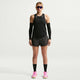 En person ser fremad, iført Nike AeroSwift Running Singlet Dame Black/White, sorte ærmer, sorte shorts, knæhøje sorte sokker, lyserøde løbesko og blå solbriller mod en almindelig hvid baggrund.