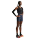 En kvinde står iført træningstøj, herunder en sort vægtvest, Nike AeroSwift Tight løbeshorts til kvinder i farverne Thunder Blue/Black/White/Linen, en sort armvarm, sorte sokker og flerfarvede løbesko, foran en hvid baggrund.