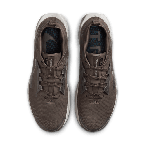 Et par Nike Pegasus Trail 5 Gore-Tex herresko i Ironstone/College Grey-Velvet Brown vises ovenfra, hvor snørebånd, mesh-struktur, hvide såler og indlægssåler med delvis branding fremhæves på en almindelig hvid baggrund.