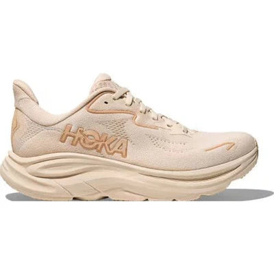 En beige Hoka-løbesko med en tyk, polstret sål og HOKA-logoet i brun farve på siden, vist i profil mod en almindelig hvid baggrund.
