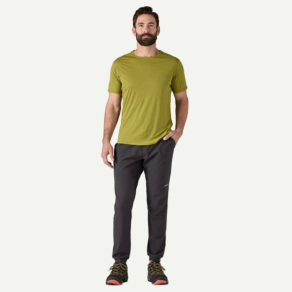 En mand med brunt hår og skæg står med front mod en almindelig hvid baggrund, iført en grøn kortærmet t-shirt, Patagonia Terrebonne Joggers Herre i sort og trail-løbesko, med den ene hånd i lommen.