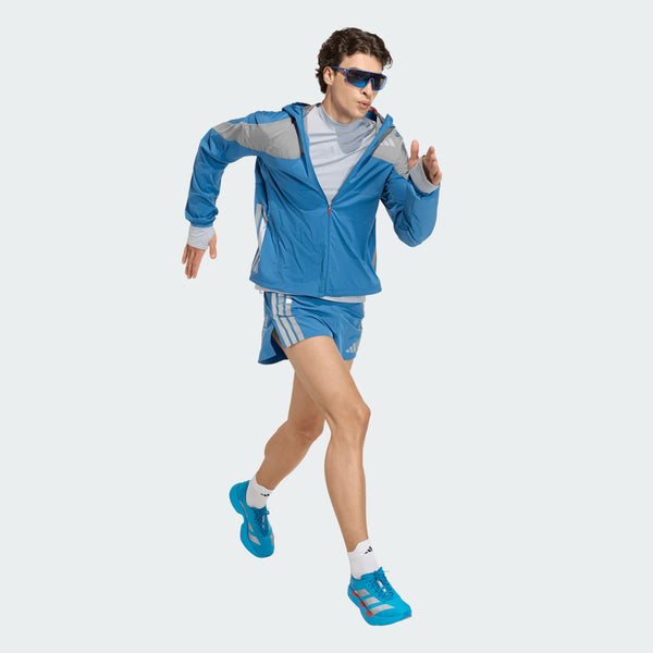 En mand iført Adidas Adizero Hakone Ekiden Jacket, Herre, Duinn/Sliver, med blå shorts, blå løbesko og solbriller løber mod en almindelig lys baggrund.