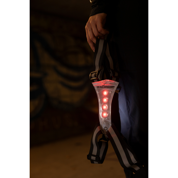 En hånd holder BeeSafe LED Harness USB Phone Pocket, Unisex, Silver, som viser fire røde lysdioder, der lyser i en lodret linje. Selen har sorte og hvide stropper og er ideel til sikre løbeture i mørke. Baggrunden er svagt oplyst og ufokuseret.