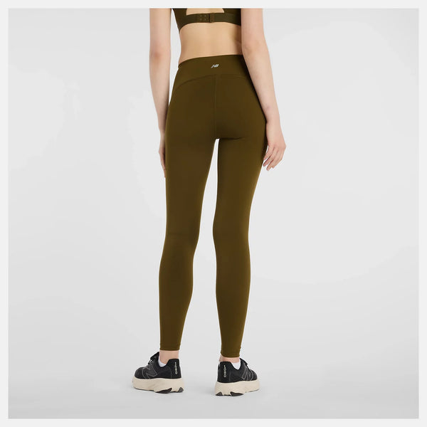 En person står væk fra kameraet i New Balance's Sleek Pocket High Rise Legging, Dame, Woodland, sammen med en matchende sports-bh og sorte sportssko med hvide såler på en almindelig hvid baggrund.