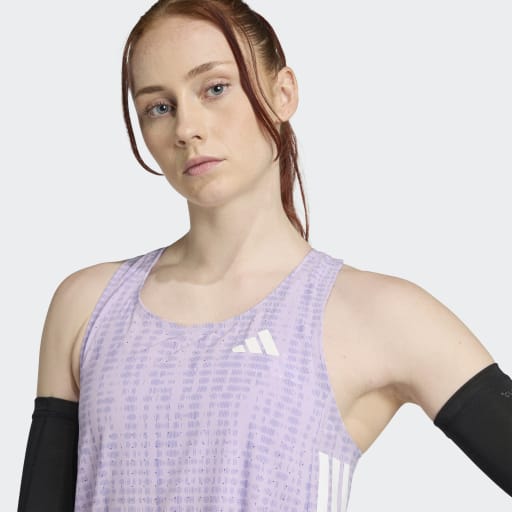En lyshåret kvinde med rødt hår er iført Adidas Adizero Singlet, Dame, Powplu/Maglil med sorte ærmer og poserer med den ene arm bøjet. Den svedtransporterende top står i kontrast til den ensfarvede, lyse baggrund.