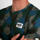 Nærbillede af en person iført Saysky Flower Combat T-Shirt, Herre, Blue Aop. Den nederste halvdel af ansigtet og overkroppen er vist, og brystmærket er fremhævet.